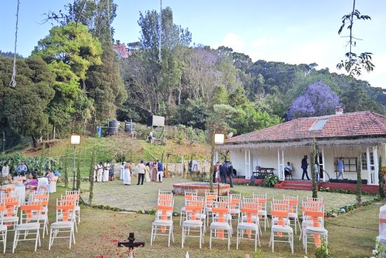 alt-text Wedding setup in Coonoor with rows of chairs and a central water feature under a tree adorned with purple flowers.