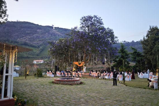 alt-text Wide shot of an outdoor wedding venue with guests seated around a central stage under a large tree with purple flowers in Coonoor.