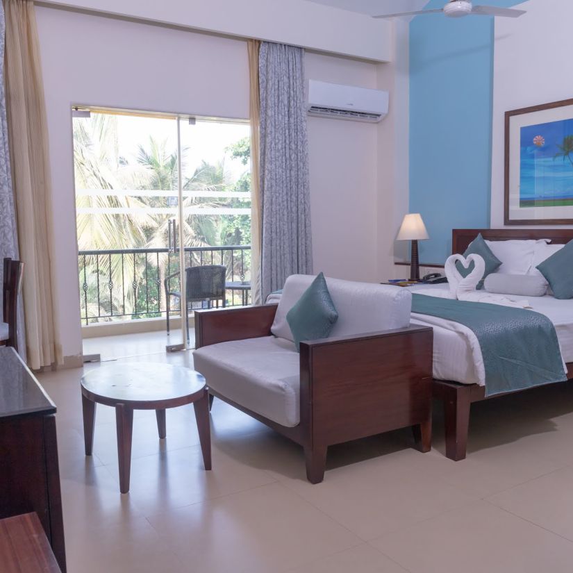 Complete view of the Deluxe room with a double bed, a couch a small tea table and a balcony at Kyriad Prestige Calangute, Goa