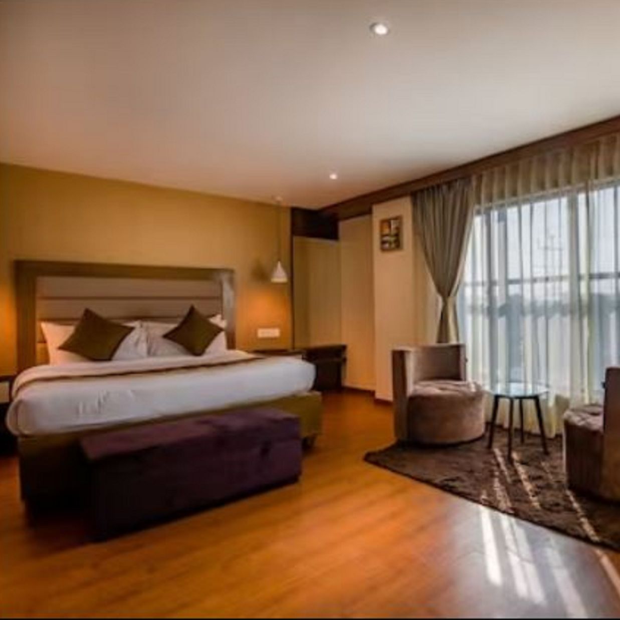 A premium hotel room with a large bed and windows that extend from the ceiling to the floor, with curtains pulled to the side, highlighting the two furniture seatings and table at Yashshree Imperial, Siliguri.