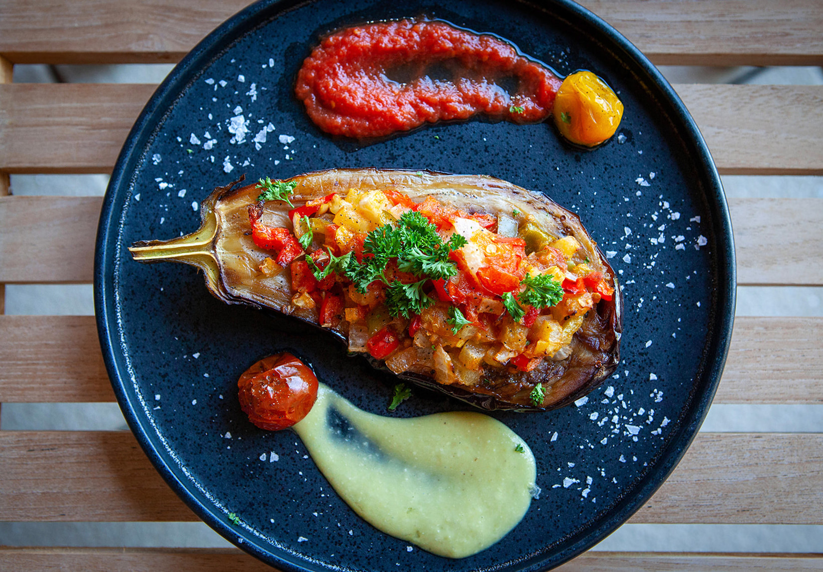 A stuffed eggplant on a black plate accompanied by a swipe of red sauce and a dollop of yellow sauce, garnished with a sprig of parsley.