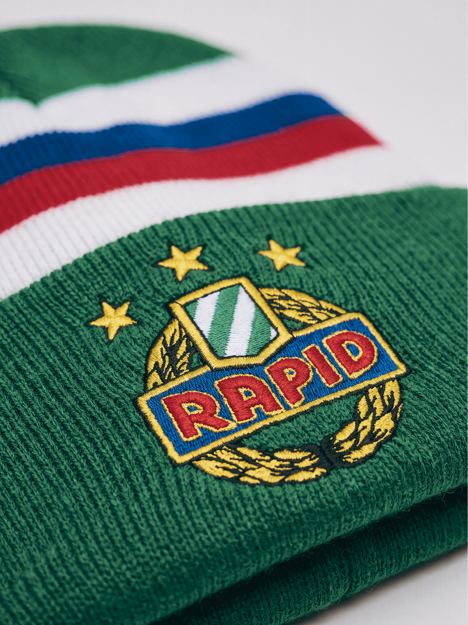 Detail der grünen SK Rapid Kinder-Strickhaube mit gesticktem Rapid-Wappen und weiß-blau-roten Streifen.