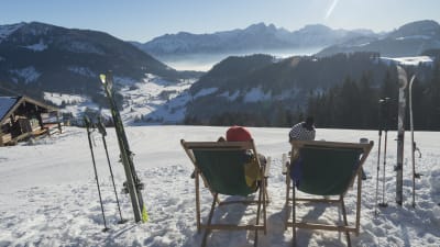 To personer siddende i liggestole med skiene i sneen og udsift over bjergene i solen