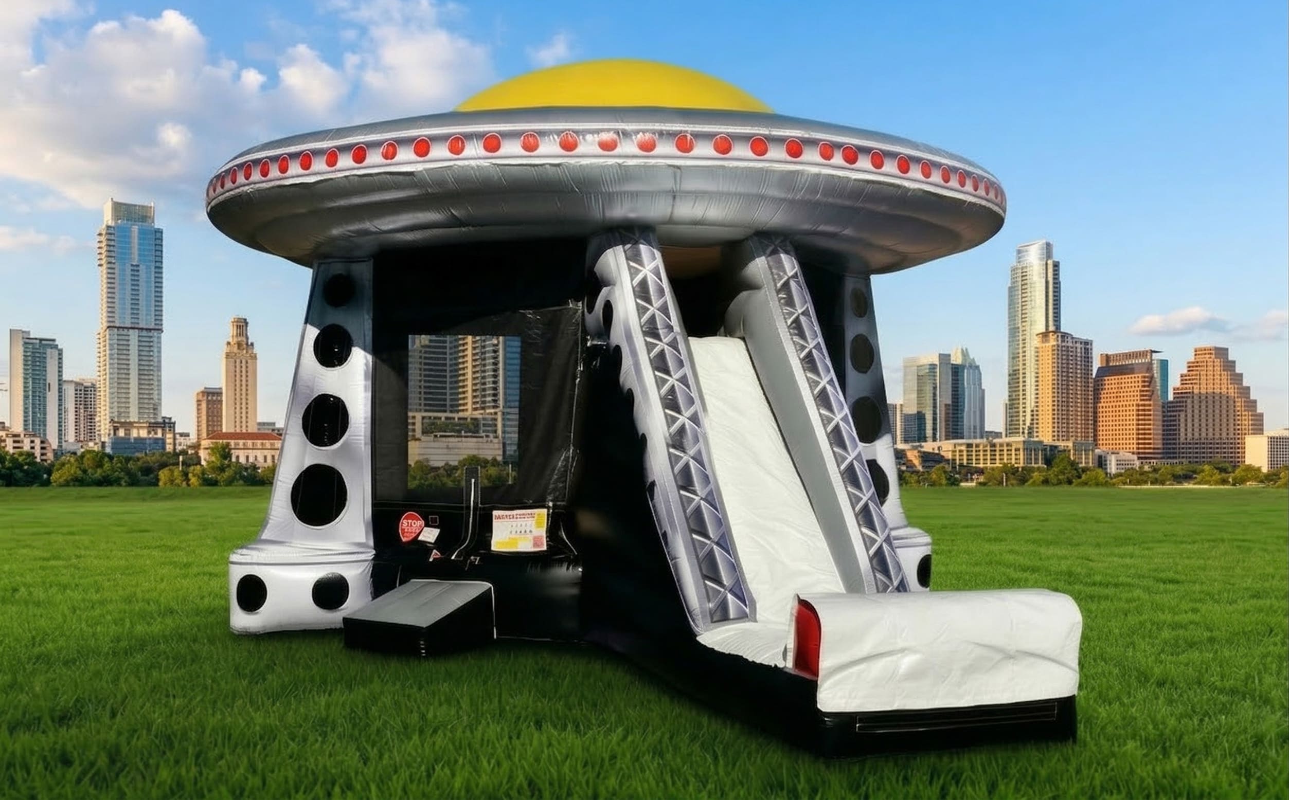 A dynamic, eye-catching image featuring a large, sci-fi-themed alien spaceship inflatable bounce house and slide setup in a sprawling urban park, with the recognizable skyline of downtown Austin, Texas, including its unique skyscrapers like the "Jenga" In