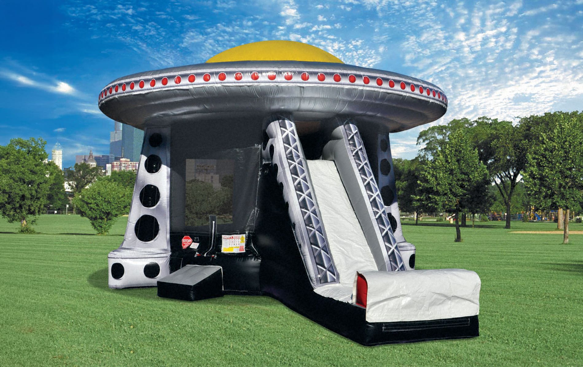 A wide, vibrant photograph of a colossal, silver and black alien spacecraft-themed inflatable bouncer set up on a massive field of green grass. The inflatable features a prominent yellow dome top, a line of flashing red "lights" around the rim, a large bl