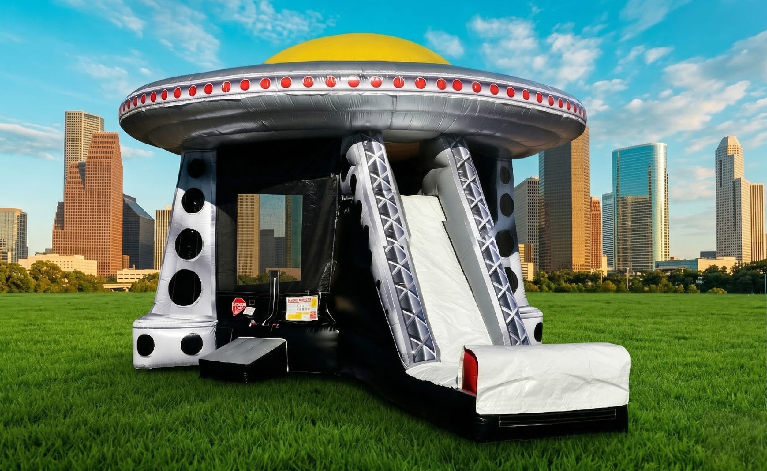 A lively photo of a massive, detailed alien saucer inflatable bounce house and slide set up in a large green park, with the distinct, modern skyline of downtown Houston visible in the distance under a bright, partly cloudy sky. The bouncer, a popular choi