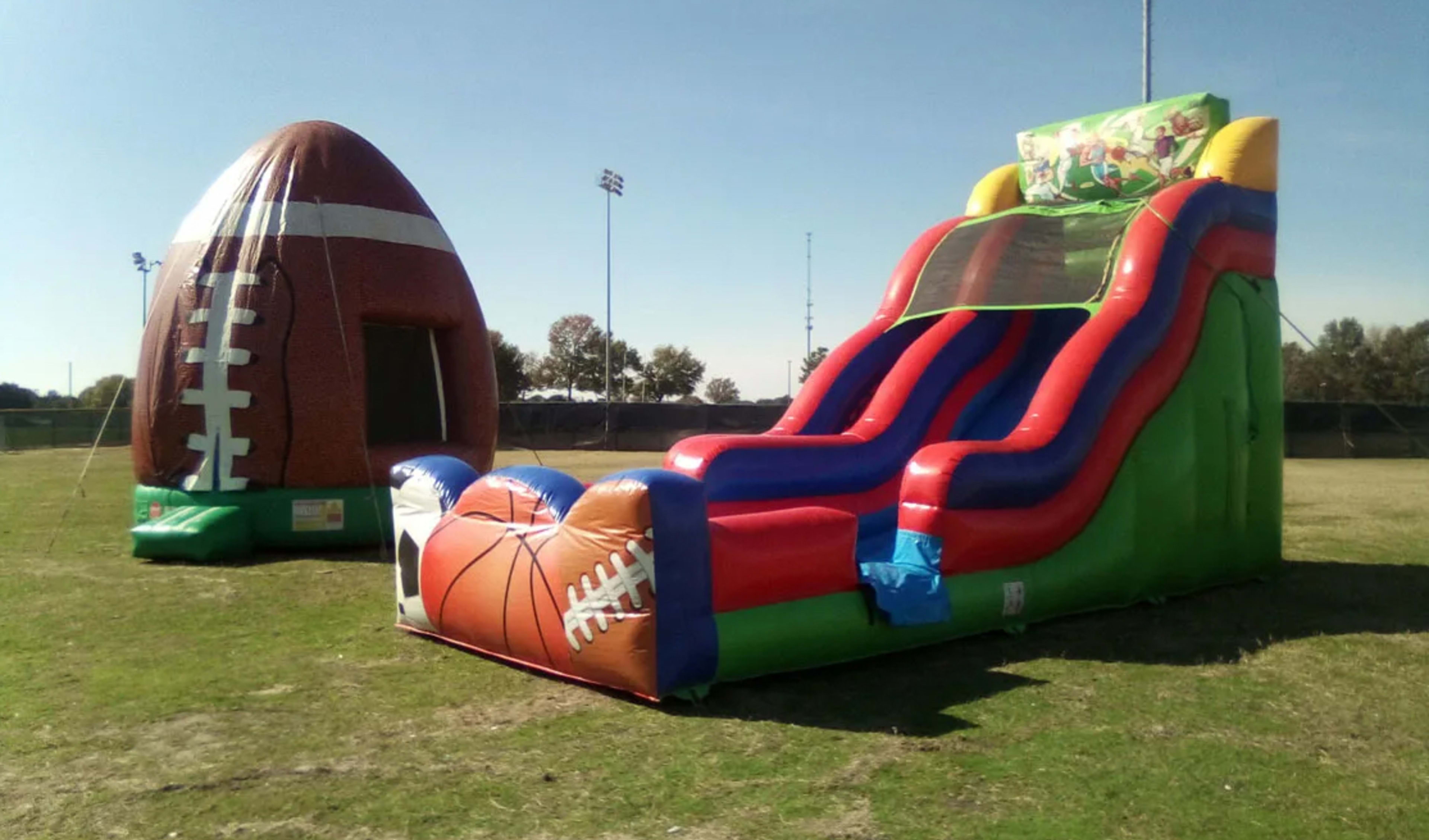 An outdoor field day setup featuring a sports-themed inflatable water slide and a large football-shaped bounce house on a grassy park area. The colorful slide features basketball and football graphics, while the separate football bouncer provides an immer