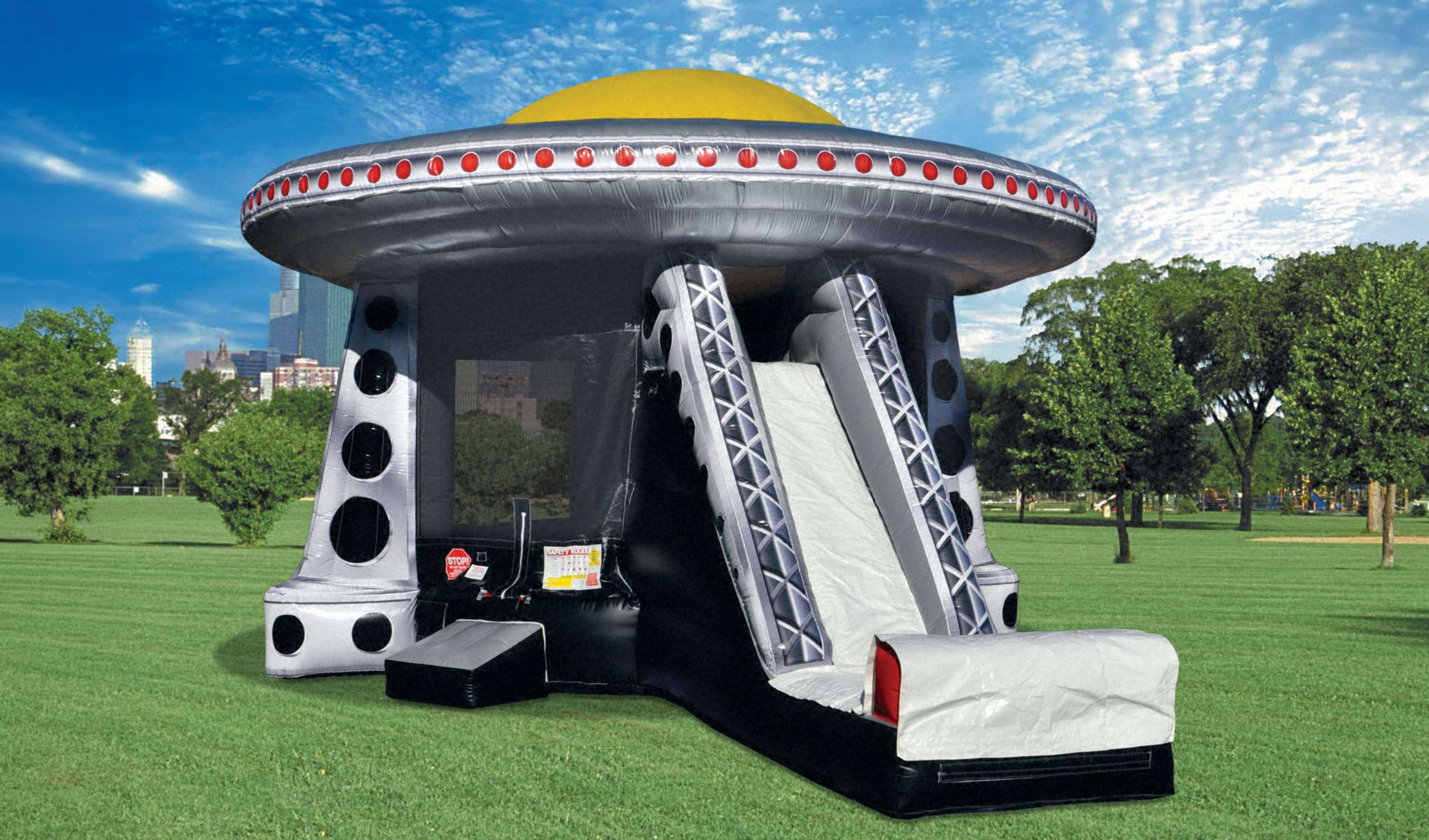 A wide, vibrant photograph of a colossal, silver and black alien spacecraft-themed inflatable bouncer set up on a massive field of green grass. The inflatable features a prominent yellow dome top, a line of flashing red "lights" around the rim, a large bl