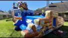 A large "Bear Camp" themed inflatable water slide and play center featuring a giant grizzly bear with a snorkel and blue wave details. The backyard scene includes families enjoying a party with popcorn and cotton candy stations, balloon bouquets, and outd Youtube