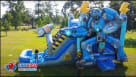 A detailed view of a blue and grey robot-themed inflatable combo unit featuring a wide climbing area and a long water slide. The backyard celebration includes families enjoying snacks from a concession stand and kids playing in a lush green park setting.  Youtube
