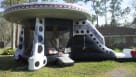 A wide, vibrant photograph of a colossal, silver and black alien spacecraft-themed inflatable bouncer set up on a massive field of green grass. The inflatable features a prominent yellow dome top, a line of flashing red "lights" around the rim, a large bl Youtube