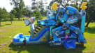 A detailed view of a blue and grey robot-themed inflatable combo unit featuring a wide climbing area and a long water slide. The backyard celebration includes families enjoying snacks from a concession stand and kids playing in a lush green park setting.  Youtube