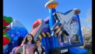 An outdoor birthday celebration featuring a space shuttle inflatable slide and moonwalk combo on a manicured lawn. The scene includes an astronaut balloon display, a themed cake table, and children playing in the water slide. Adults are shown supervising  Youtube
