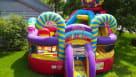 A large, candy-themed inflatable play center featuring giant lollipops, candy cane arches, and a gumball machine topper. The backyard scene shows children playing in the inflatable while families enjoy a party with popcorn, cotton candy, and snow cone sta Youtube