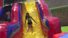 A vibrant backyard summer party featuring a large "Sports Splash" inflatable water slide with a football scoreboard topper. Multi-ethnic children are sliding and playing near basketball and soccer ball-themed landing walls. The scene includes adults and k Youtube