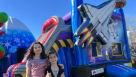 An outdoor birthday celebration featuring a space shuttle inflatable slide and moonwalk combo on a manicured lawn. The scene includes an astronaut balloon display, a themed cake table, and children playing in the water slide. Adults are shown supervising  Youtube
