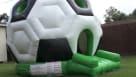 A massive black and white soccer ball-shaped moonwalk rental set up on a green lawn. The scene shows a complete party package with child-sized seating, a popcorn machine, and a cotton candy station. Adults are seated nearby under a clear sky. We provide f Youtube