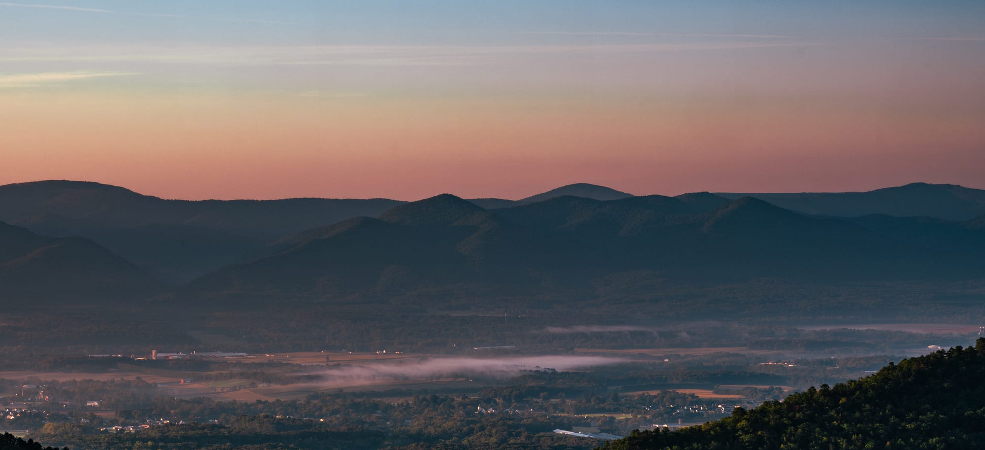 A dawn valley under a graded sky — deep blue overhead easing to ember at the horizon, a small town waking below.