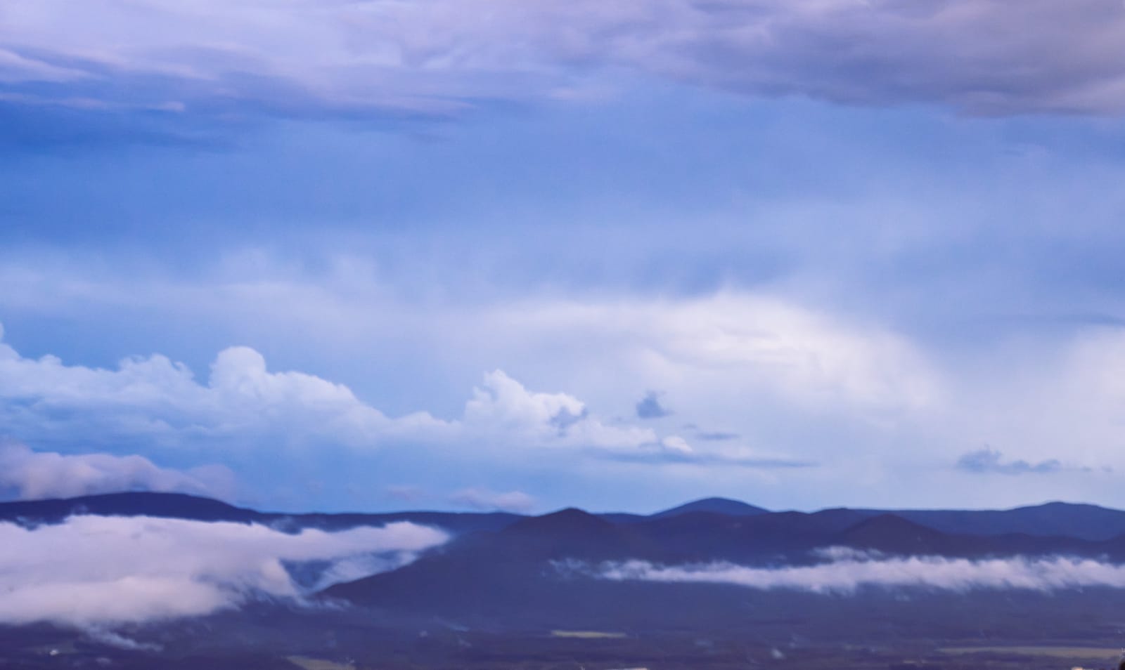 Layered dark mountain ridges partially veiled by ribbons of white fog beneath an expansive blue and lavender sky.