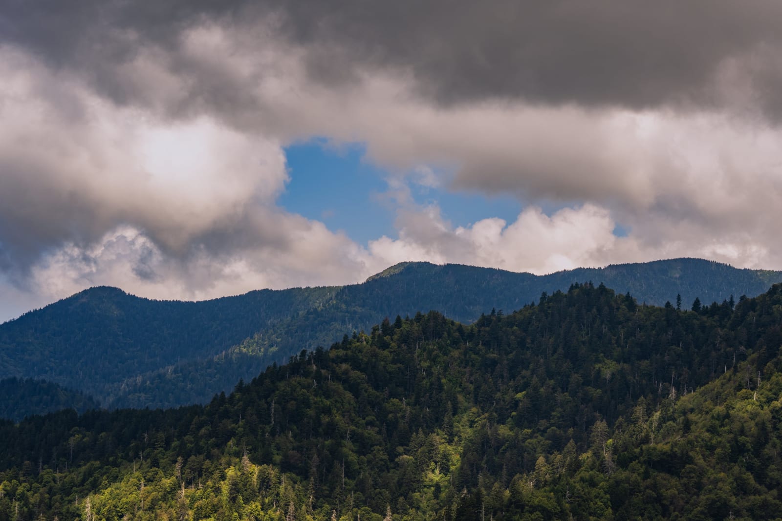 Layered forested mountain ridges beneath dramatic storm clouds, with sunlight breaking through a vivid opening of blue sky.