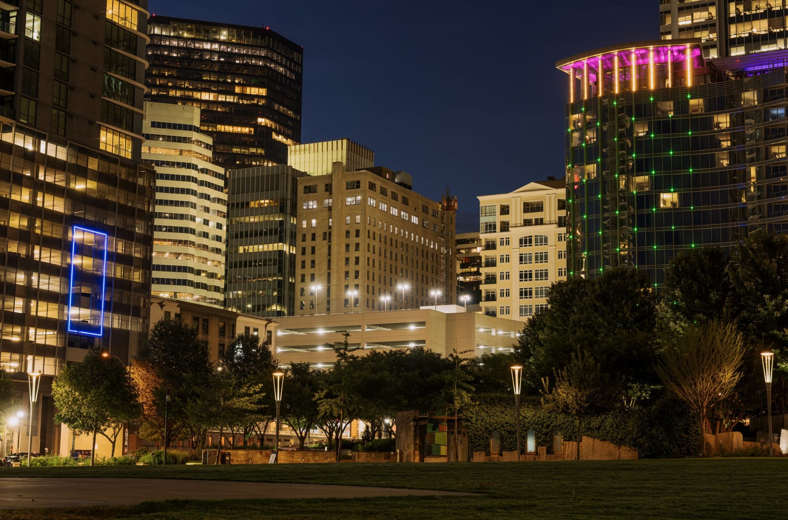 A night view of the glowing Uptown Charlotte, North Carolina skyline rising behind the warm park lanterns and green lawn of Romare Bearden Park.