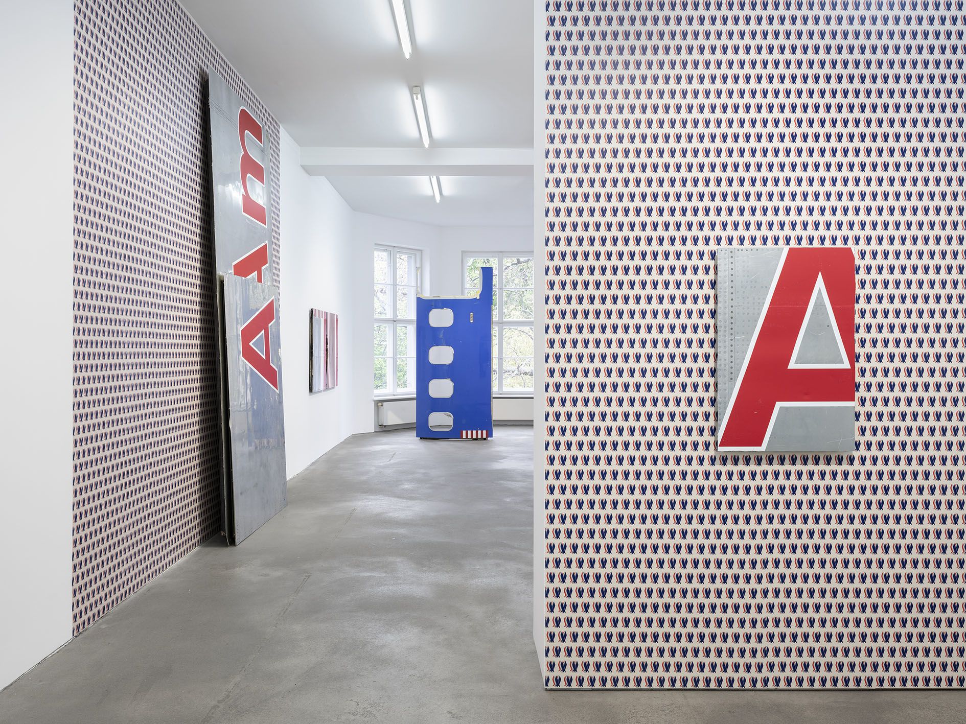 Art gallery exhibition space showing walls with patterned wallpaper featuring small blue and red motifs, large red letter "A" displays, and a clear view down the corridor