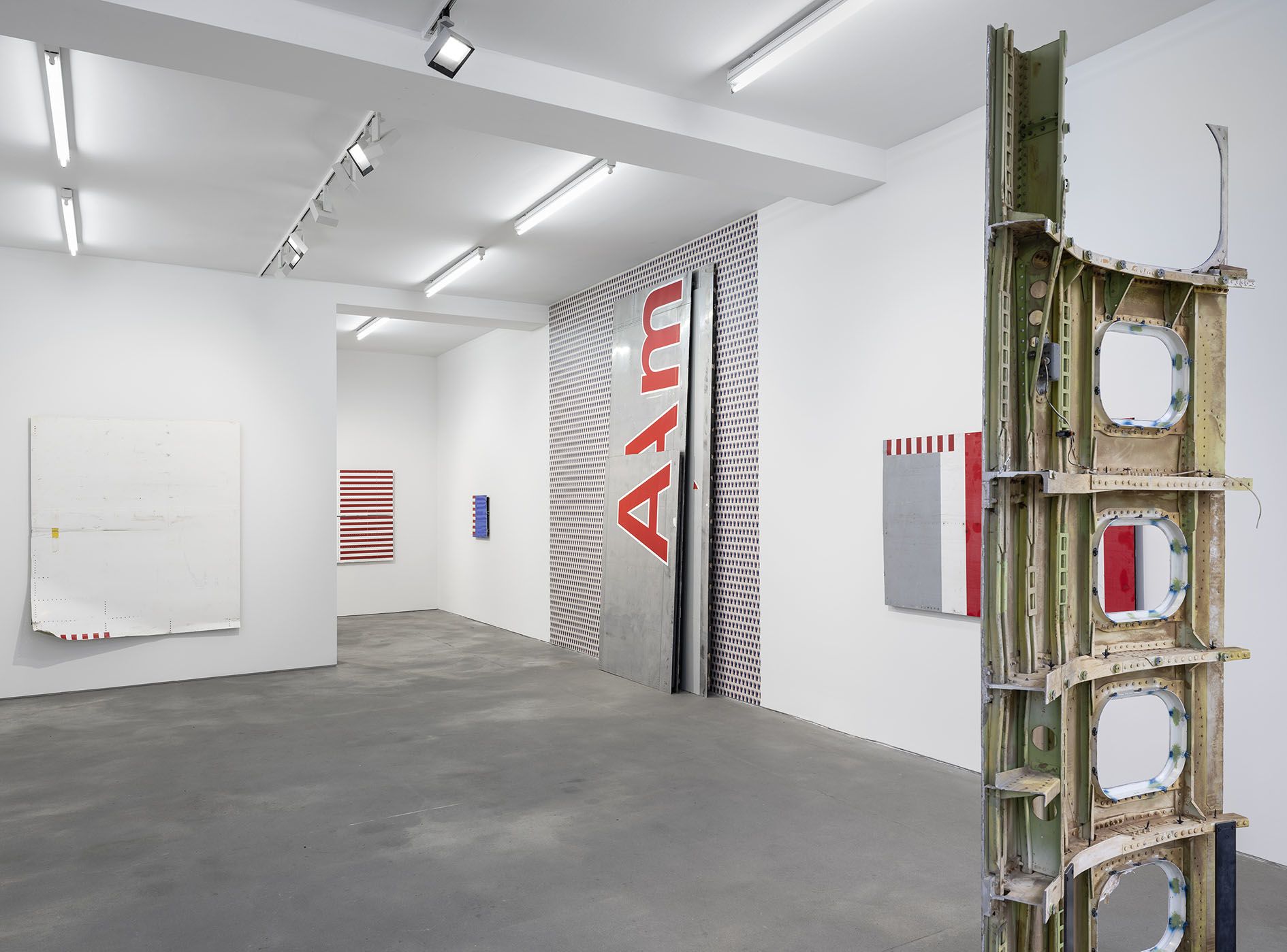 Gallery installation showing various artworks including an aircraft fuselage with windows on the right and striped red and white panels on the walls