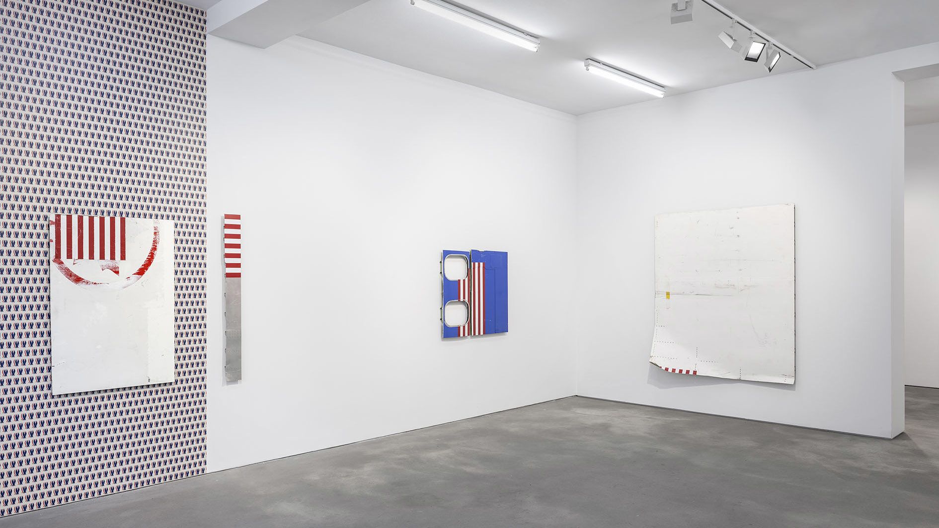 Contemporary art gallery with white walls displaying several minimalist artworks featuring red and white stripes, blue panels, and a distinctive patterned wall with small repeated motifs