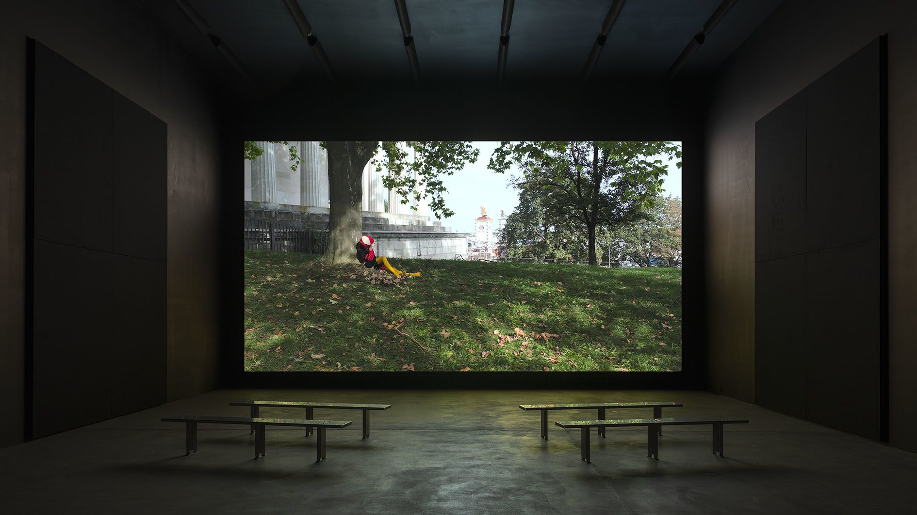 Gallery space with benches facing a large projection screen displaying a park scene with a person on grass near classical columns.