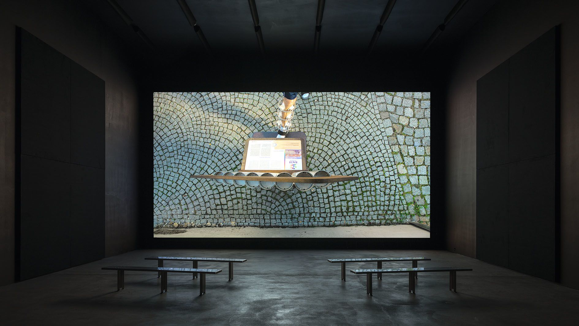 Gallery installation with benches facing a large screen showing mosaic tilework and what appears to be a small altar or shrine setup.