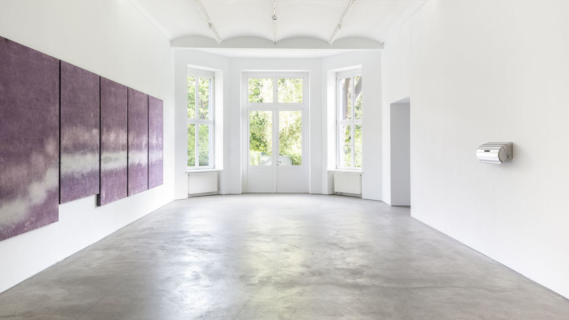 Modern gallery space with purple abstract paintings on white walls, polished concrete floors, and large windows overlooking greenery.