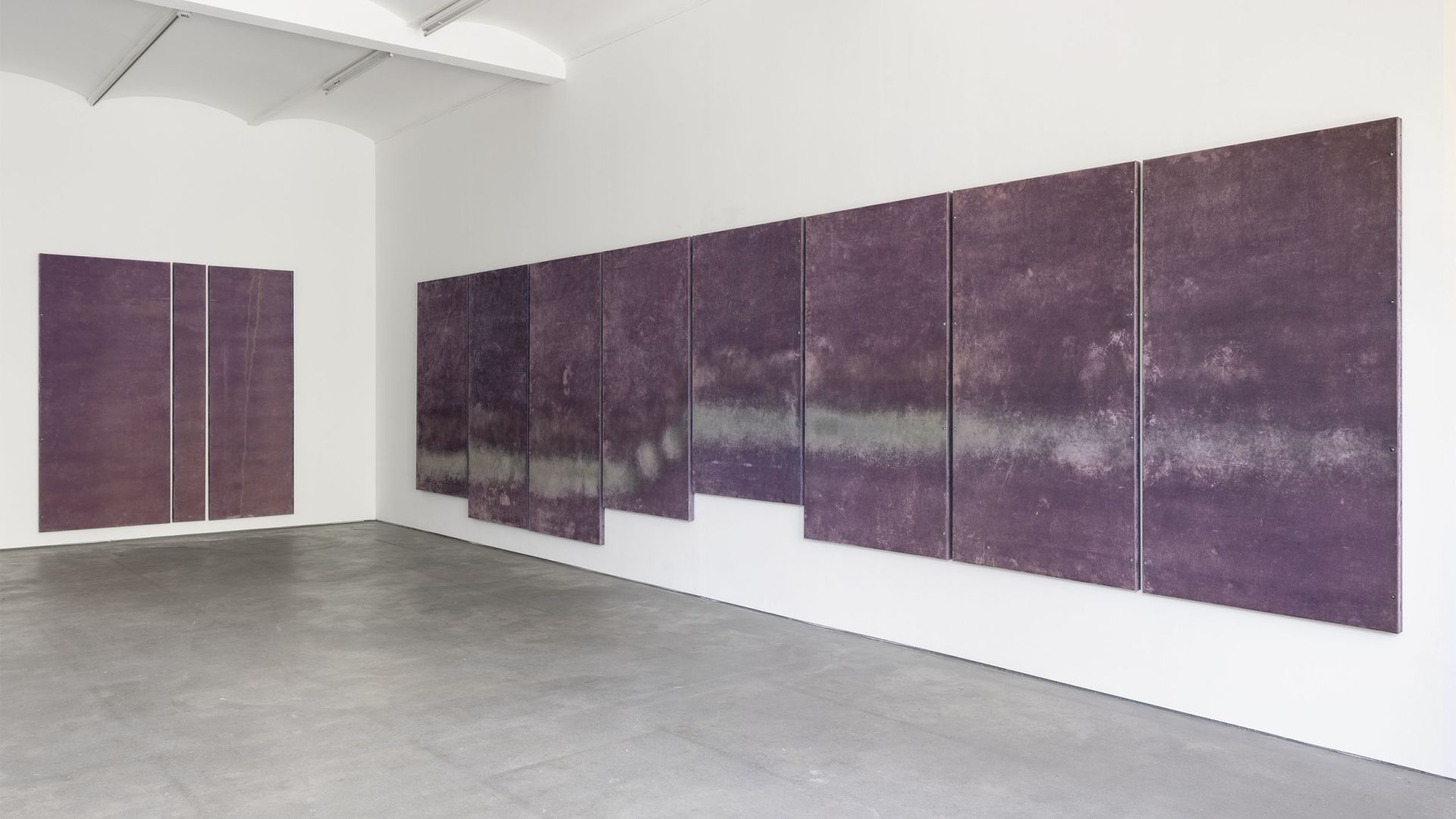 Gallery corner view showing purple abstract paintings on two walls with polished concrete floor and track lighting above.