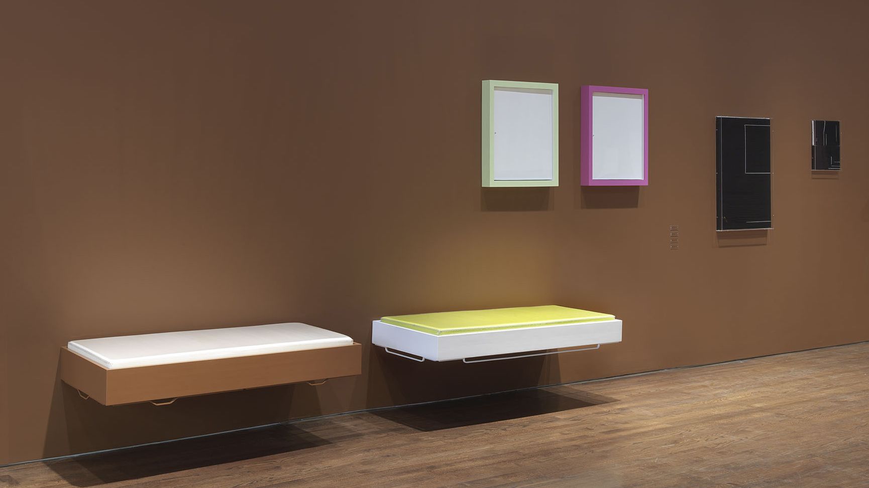 Gallery installation with warm brown walls featuring two wall-mounted rectangular frames with pale green and pink borders, plus two floating benches with white cushions, one with a wooden frame and one with a white frame.