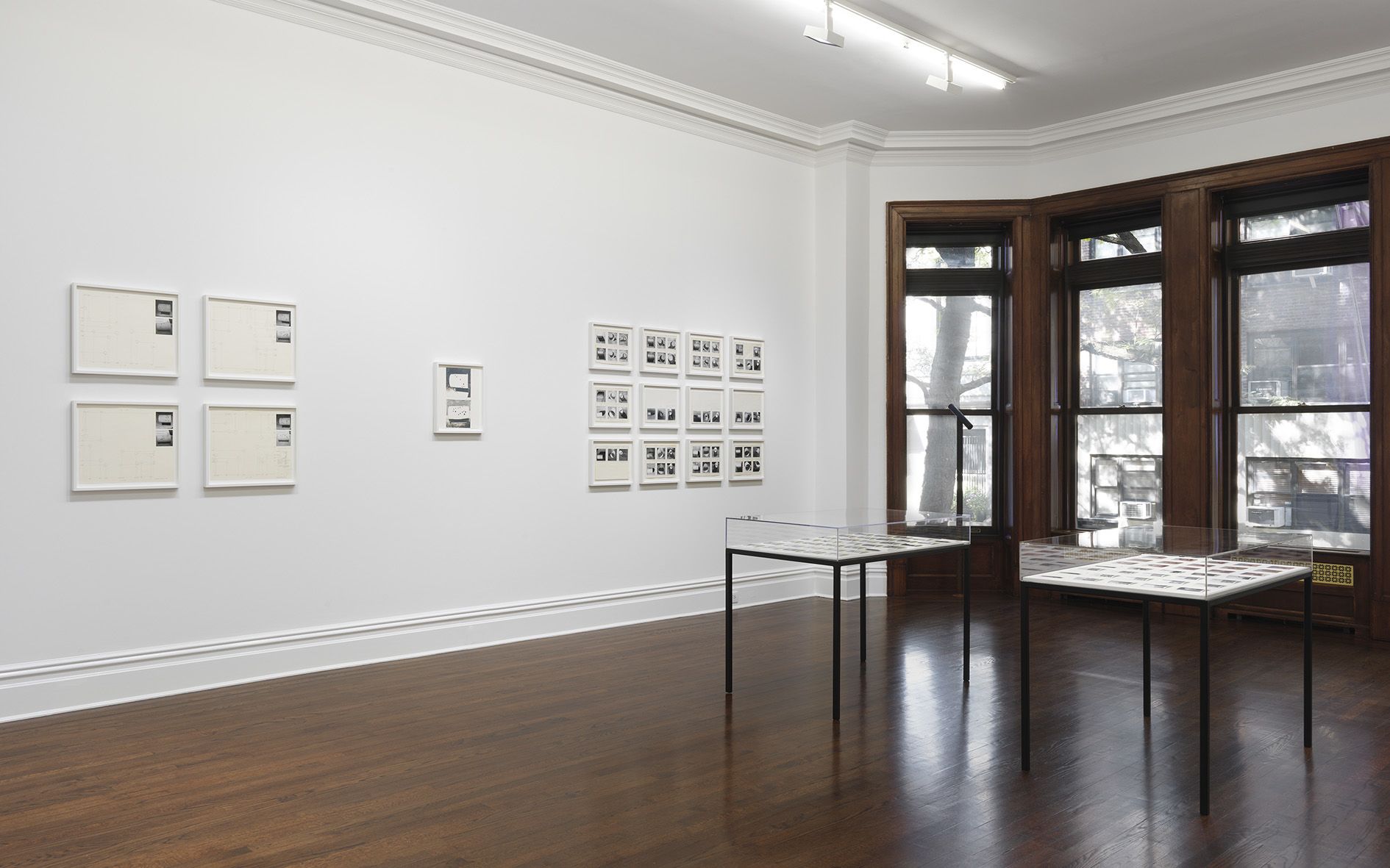 Gallery view showing archival materials in glass display cases and framed photographic works arranged on white walls with natural window lighting.