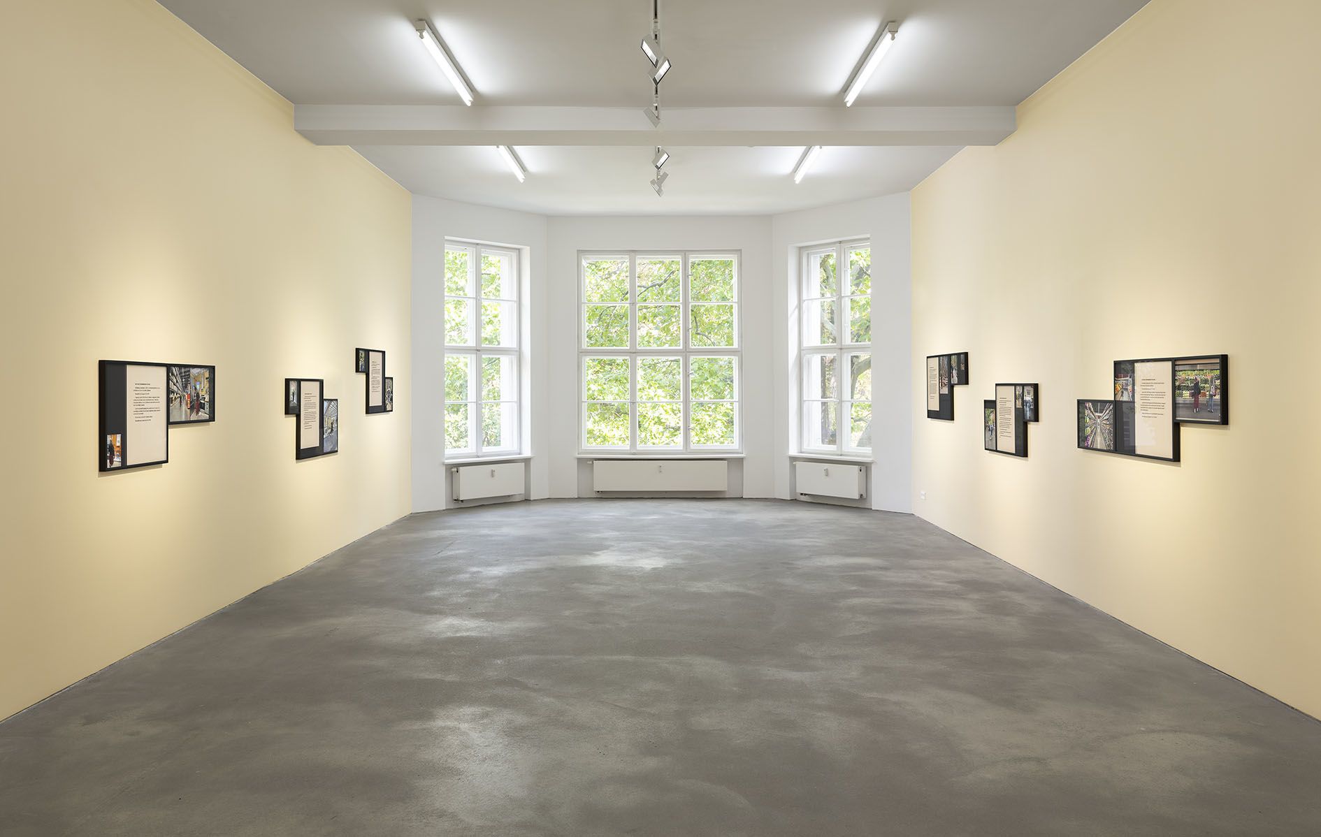 Gallery space with cream walls showing multiple framed artworks arranged in clusters, polished concrete floor, exposed ceiling beams with track lighting.