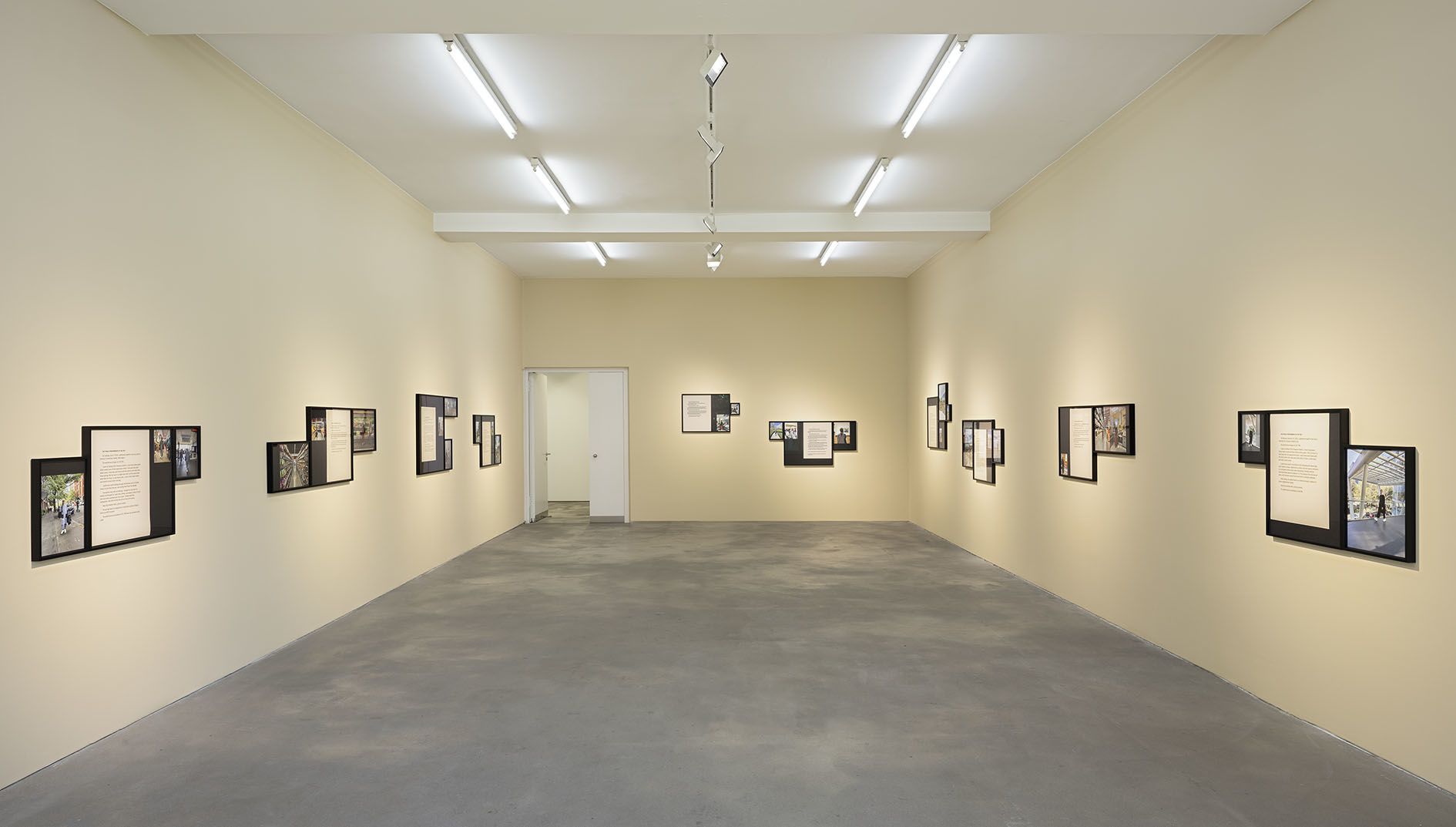 Full gallery view showing complete exhibition installation with multiple framed works systematically arranged around white-walled space with concrete floors.