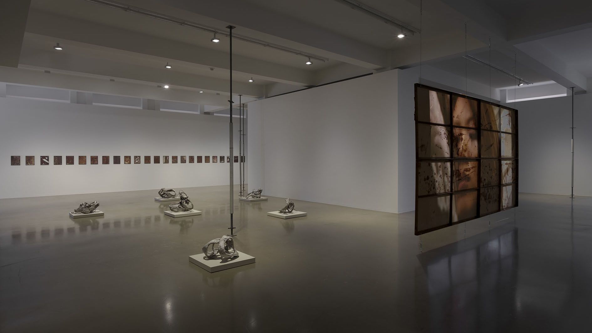 Gallery overview showing row of small artworks on wall, sculptural pieces on pedestals, and large grid artwork.