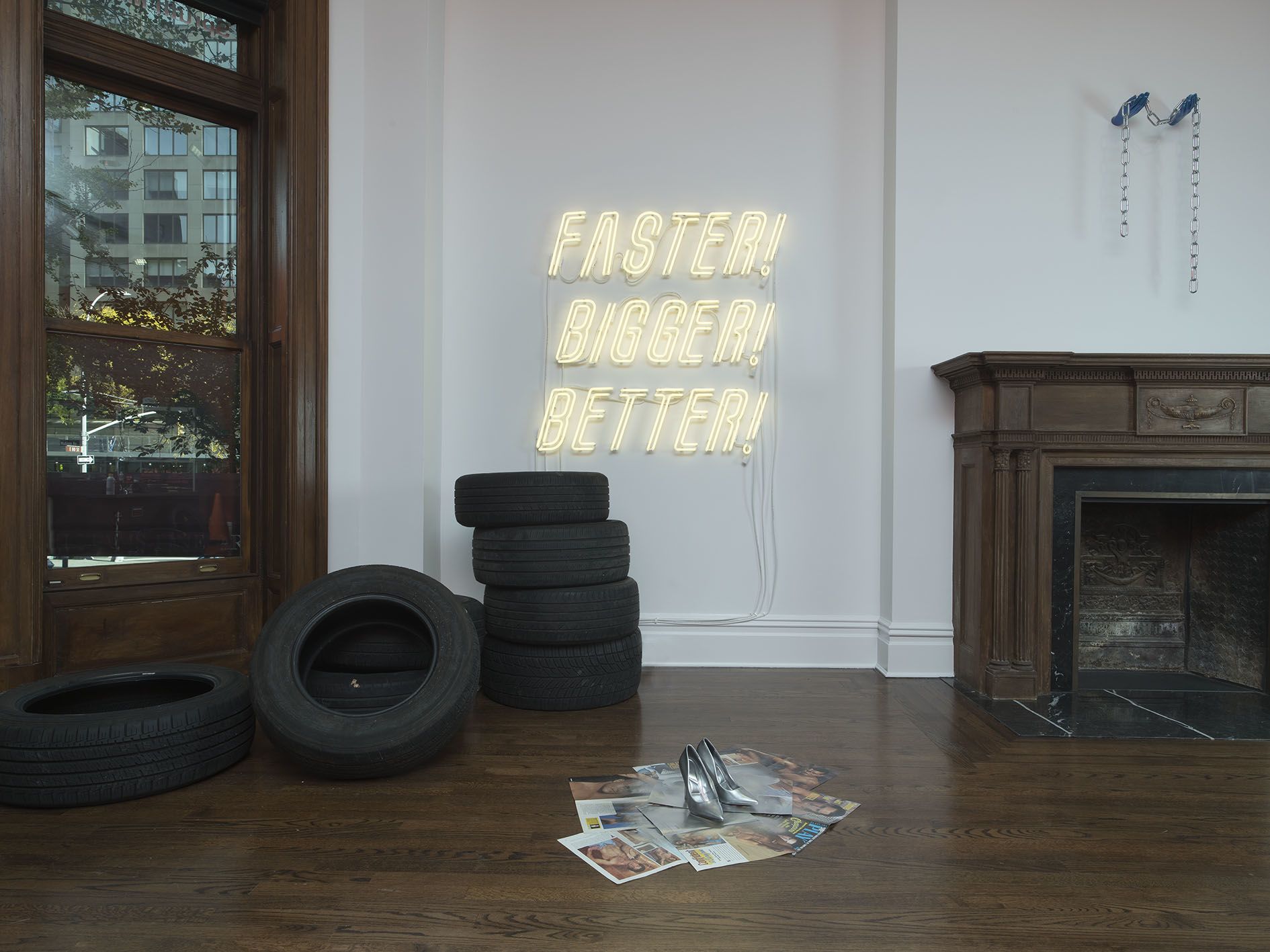 Room with yellow neon "FASTER! BIGGER! BETTER!" sign above stacked tires and scattered photos with silver shoes.