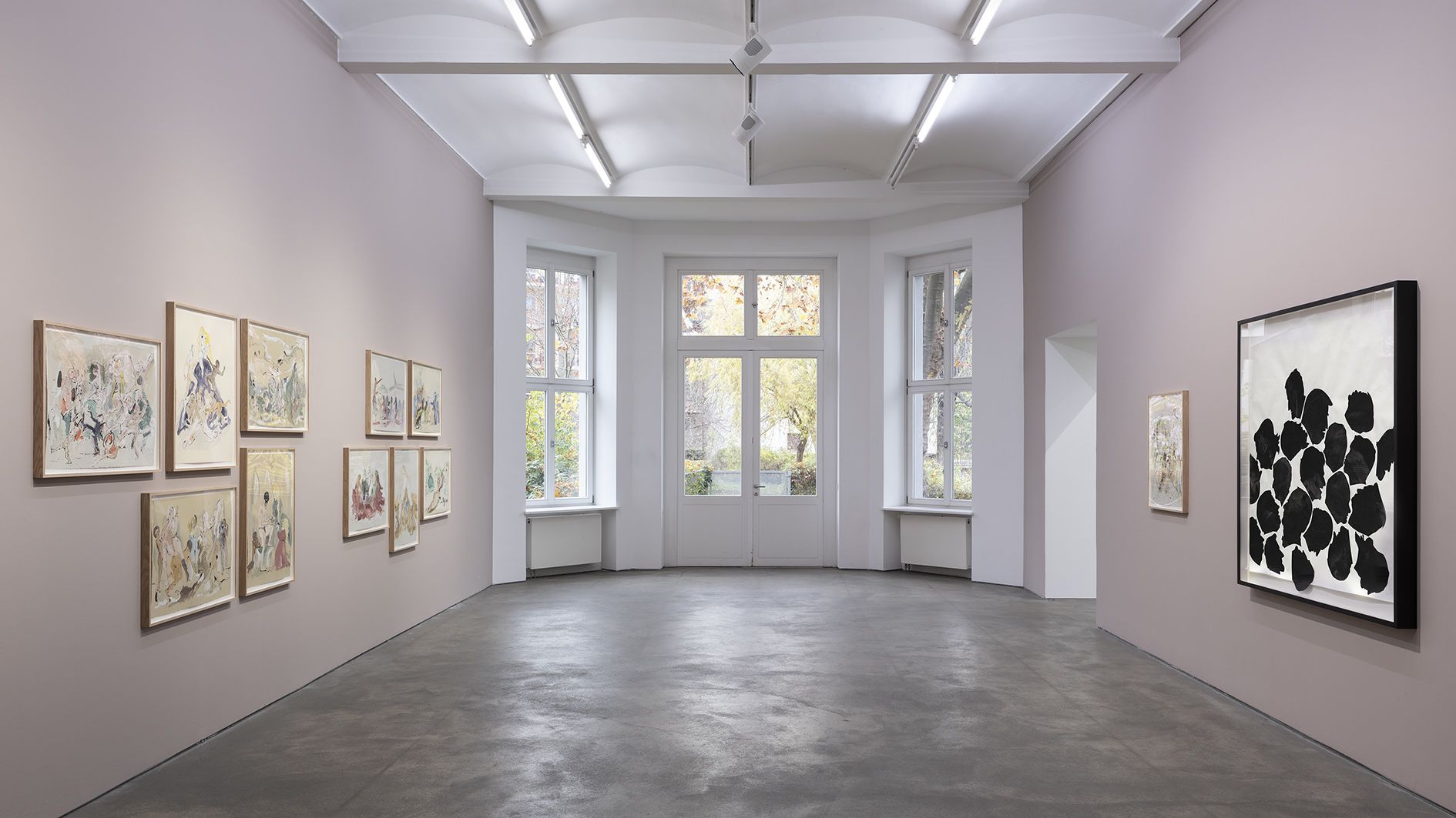 Light pink gallery space with wooden-framed artworks on left wall, silhouette piece on right, and large windows showing autumn foliage.