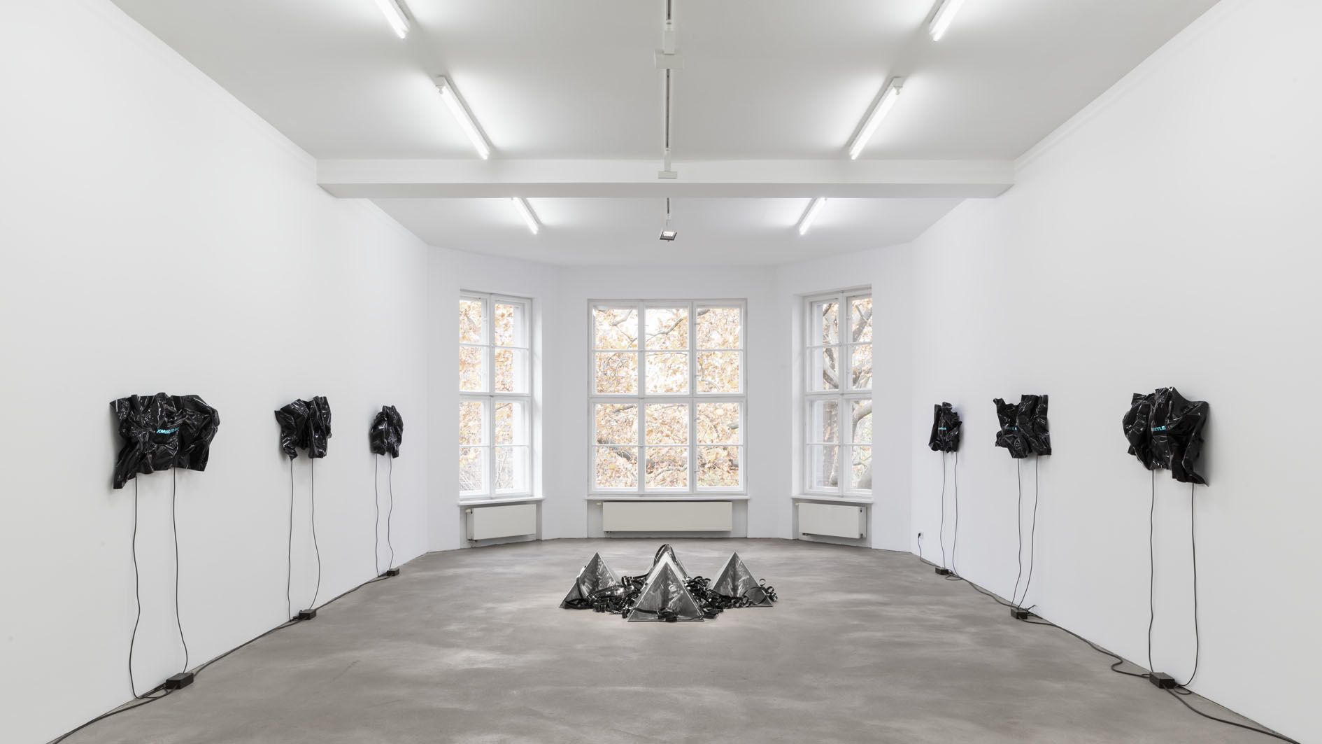 Gallery installation with seven heat-folded black vinyl sheets on white walls and metal pyramids on floor center.