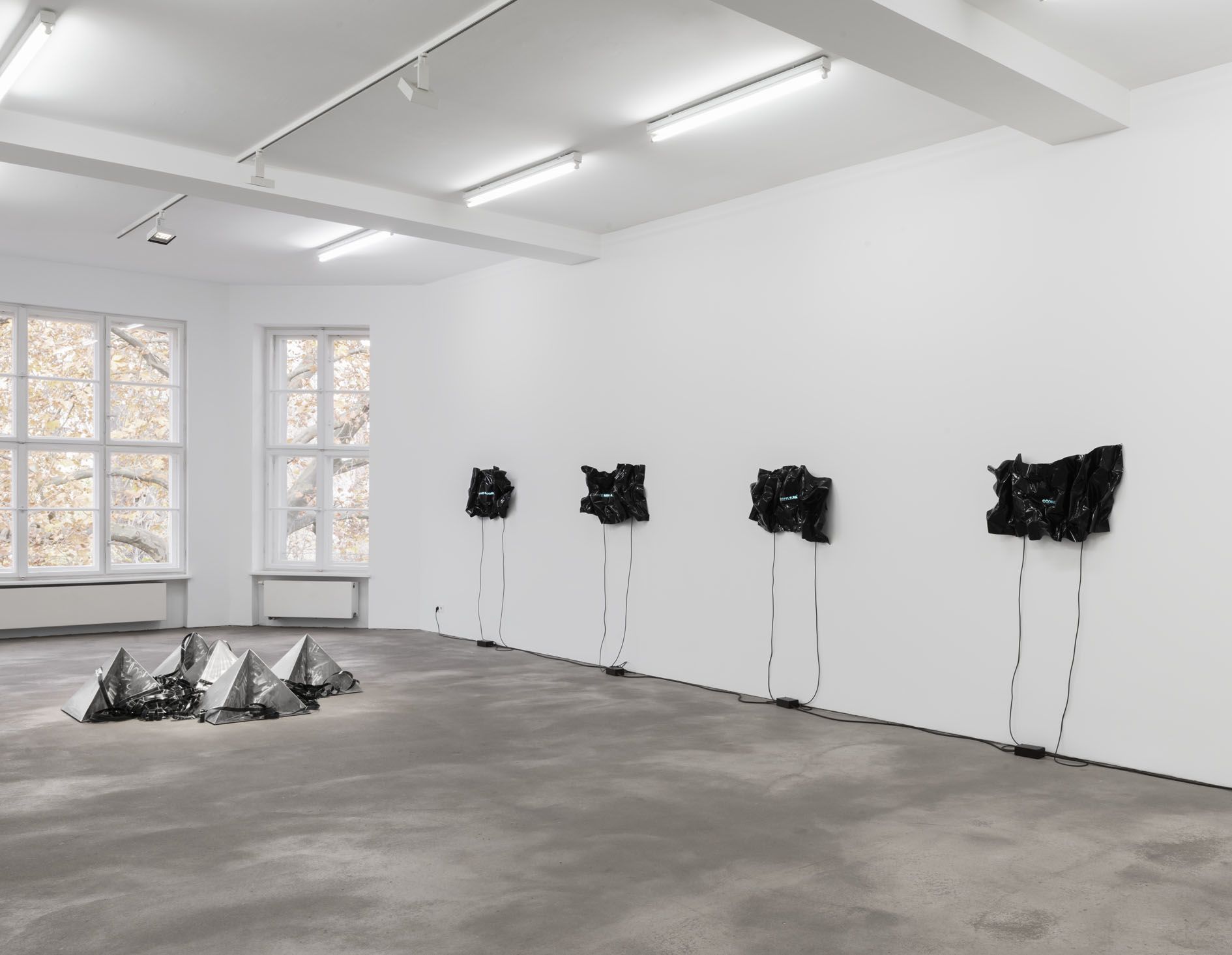 Gallery installation with four black heat-folded vinyl sheets on white walls, each displaying blue neon movie titles and reflective pyramids on floor.