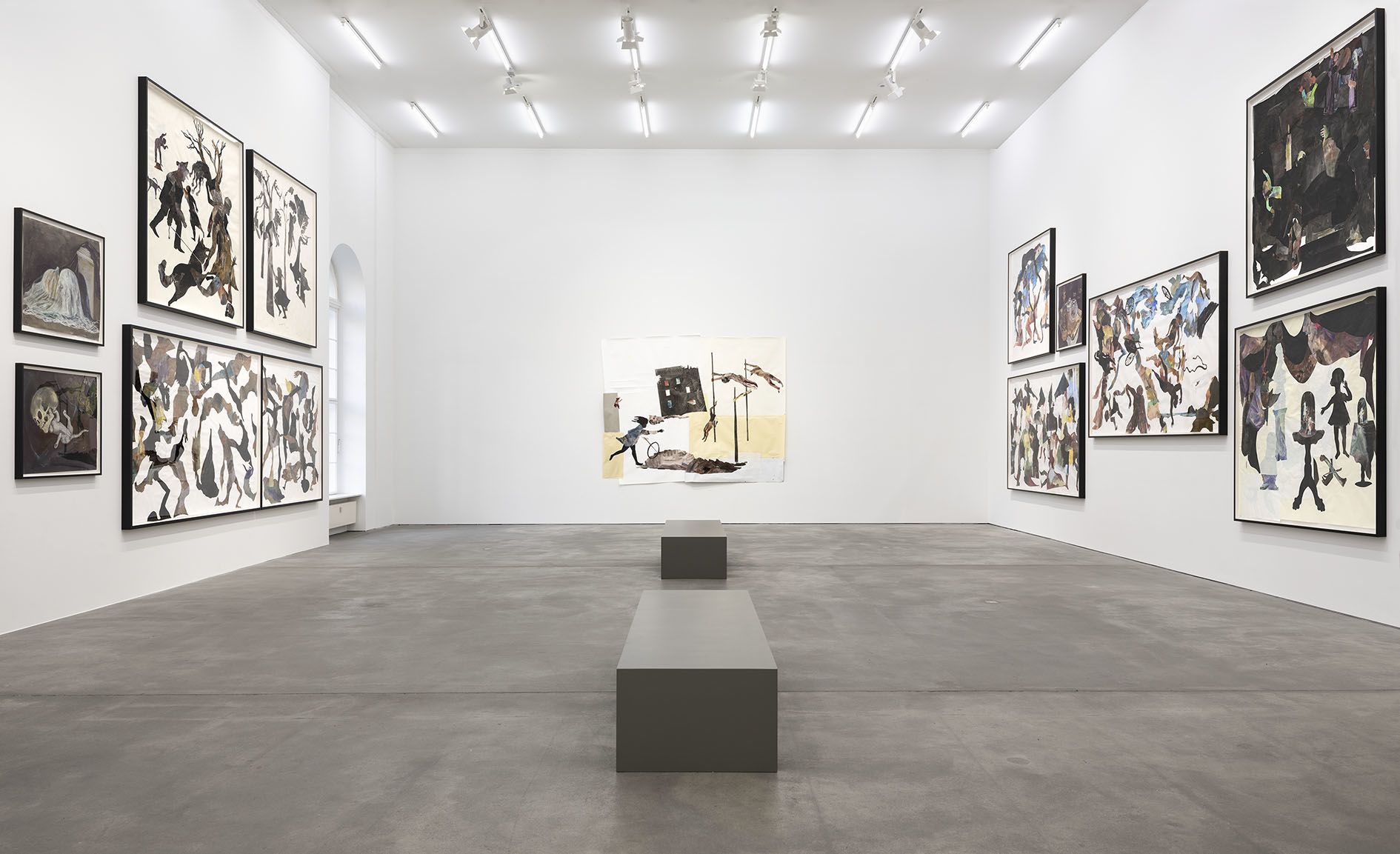 Modern gallery space with white walls, gray floor, and dark-framed artwork displayed on walls and pedestals in center.