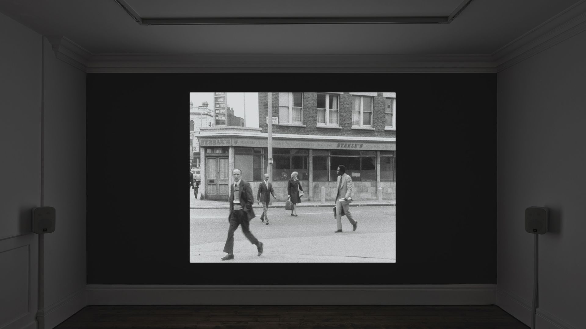 Black and white photograph of people crossing a street in front of storefronts displayed on a wall-mounted screen in a dark room