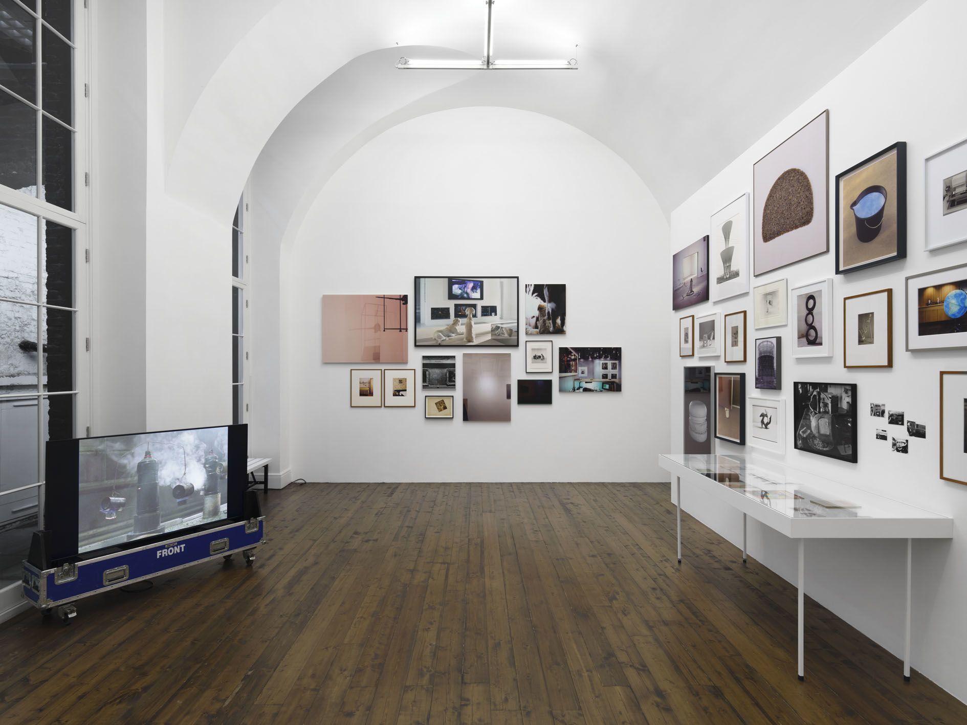 Gallery wall displaying collection of framed photographs in various sizes with video monitor on left and display case on right