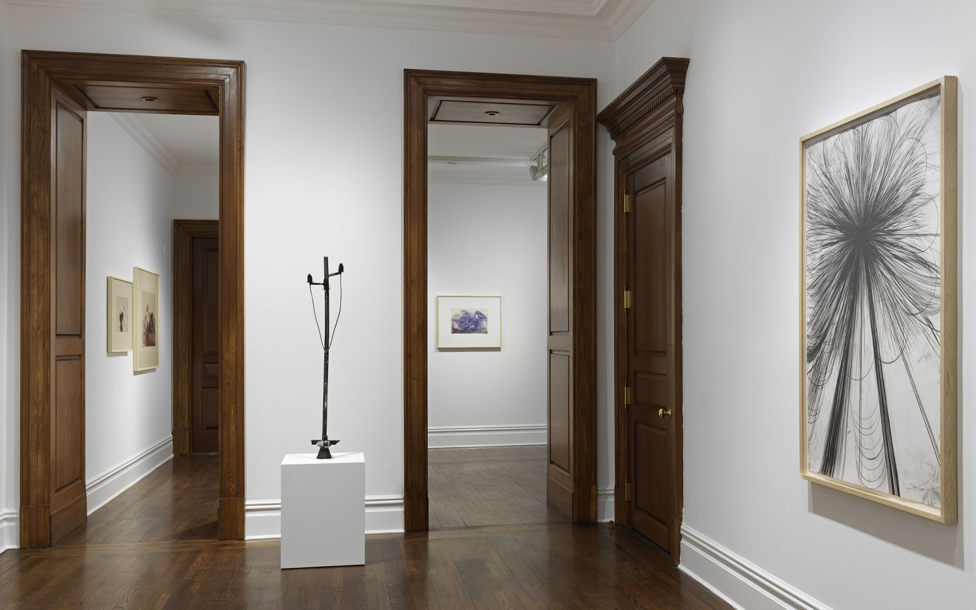 Gallery view through doorways showing framed artwork and sculpture on pedestal with wooden details