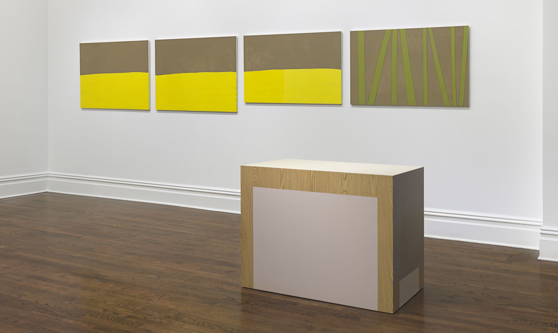 Gallery room with four brown-and-yellow abstract panels and a wood-veneer cube sculpture with grey inset panel.