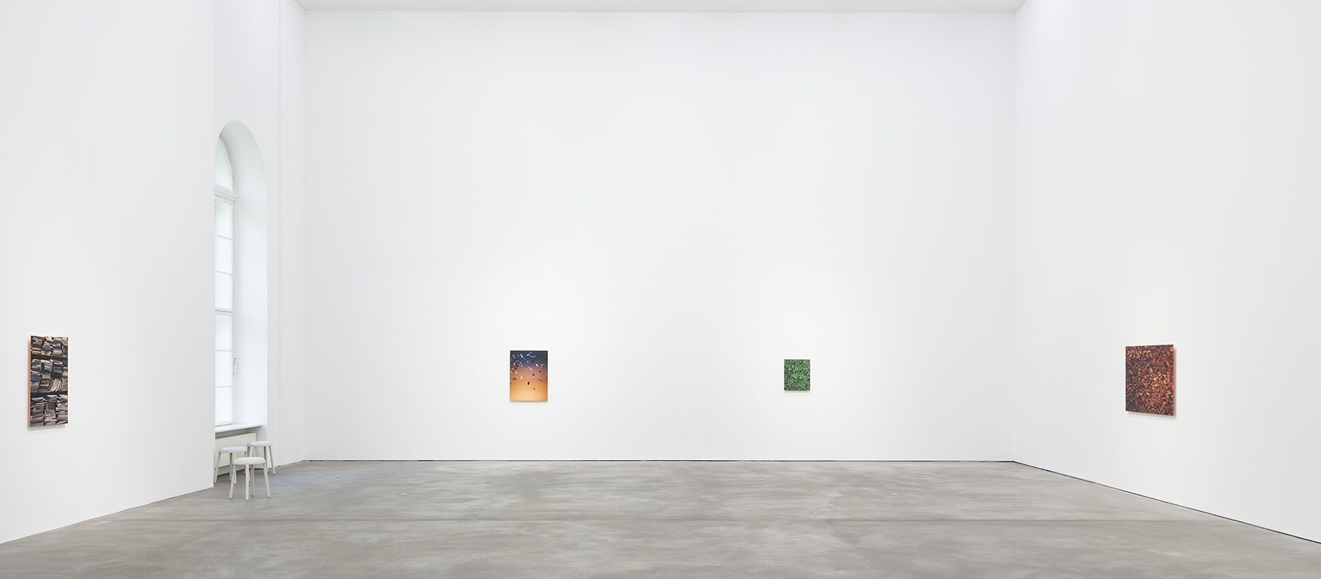 Gallery interior with four small photographs on white walls, concrete floor, arched window, white stool.