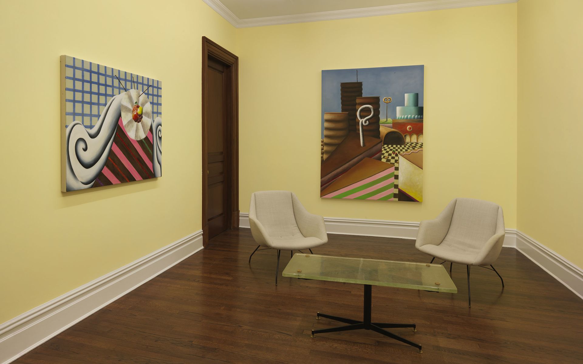 Gallery room with two large cake paintings on yellow walls; seating area with two armchairs and a glass coffee table.