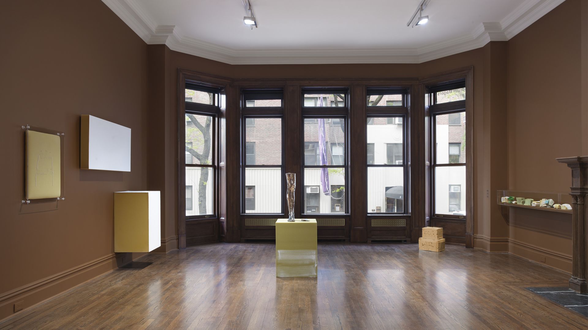 Gallery room with brown walls, wooden floors, and bay windows. Features minimalist exhibition with freestanding sculptures and wall-mounted artworks.