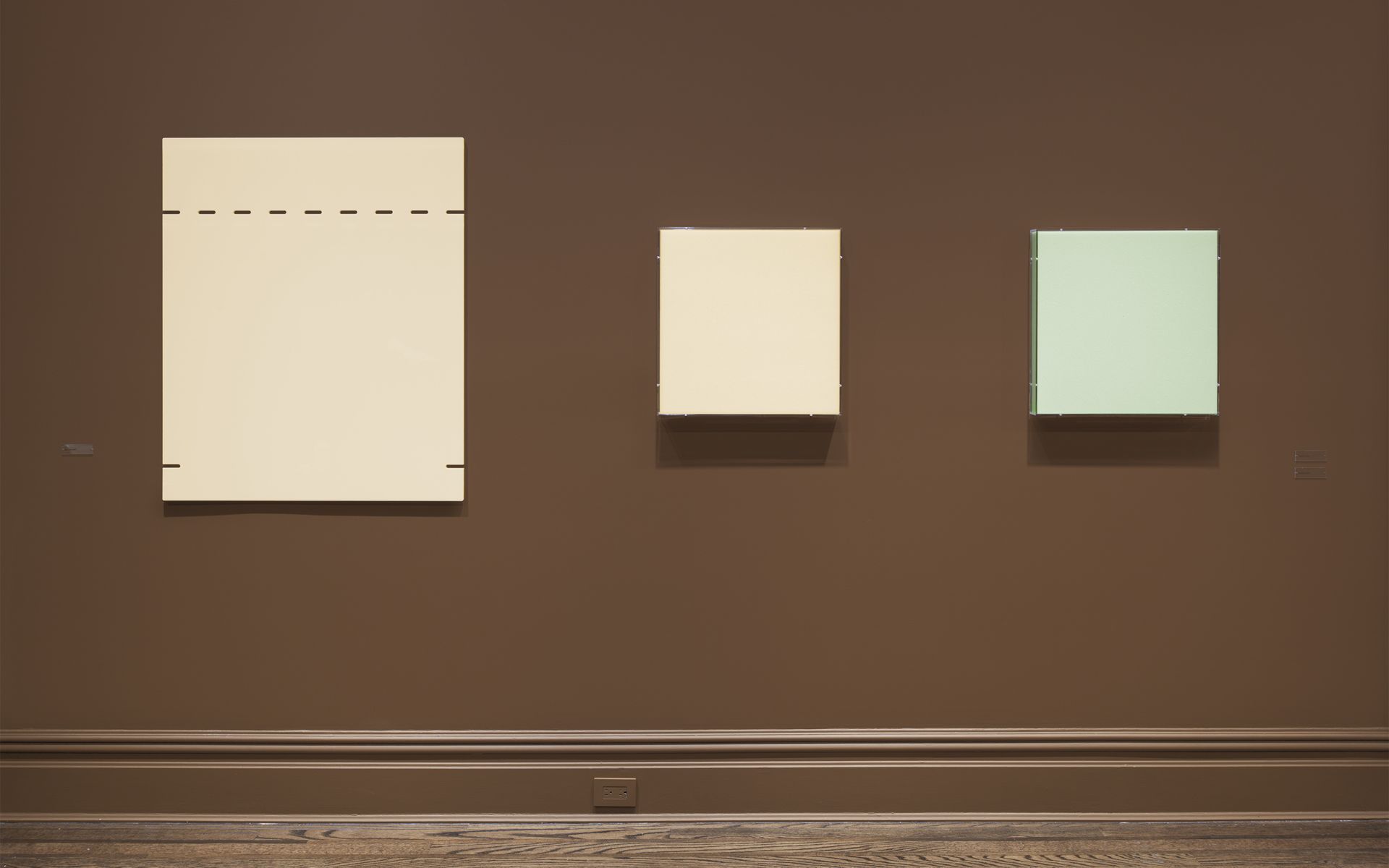 Three minimalist artworks mounted on brown gallery wall: large cream panel with dotted line and two smaller panels in cream and pale green.